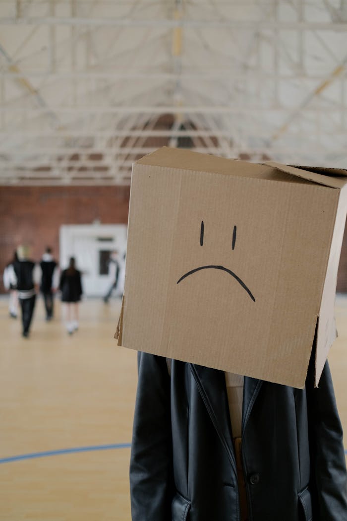 Person wearing a cardboard box with a sad emoji head in an indoor gym setting.